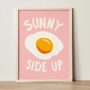 Peut inclure: Une affiche encadrée avec un cadre en bois clair. L'œuvre présente une illustration d'œuf au plat avec un jaune d'œuf orange vif et un œuf blanc sur fond rose. Les mots "SUNNY SIDE UP" sont écrits en blanc.
