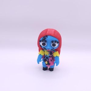 May include: A small figurine of Sally from The Nightmare Before Christmas. The figure has blue skin, red hair, and a patchwork dress with pink, black, and purple accents. Her arms and hands are blue, and she wears black shoes.