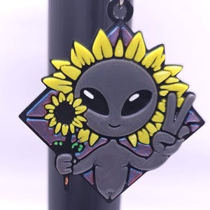 May include: A gray alien-themed keychain with a yellow sunflower design. The alien is holding a sunflower and making a peace sign. The keychain is diamond-shaped with a black border and a silver chain.