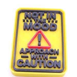 May include: A yellow sign with black text that reads "NOT IN THE MOOD" above a red triangle with an exclamation point, and "APPROACH WITH CAUTION" below. The sign has a black border.