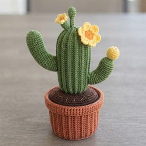 May include: A crocheted cactus in a terracotta-coloured pot. The cactus is green with vertical ridges, two side arms, and two yellow flowers with orange centres. The top of the soil is dark brown with stitched details, and the pot has a ribbed texture.