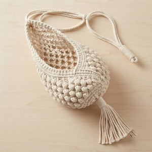May include: A cream-colored macrame hanging planter featuring a teardrop-shaped basket with a textured, knotted pattern. The upper section has an open-weave grid, while the lower body displays dense, rounded knots, finished with a long, straight tassel.