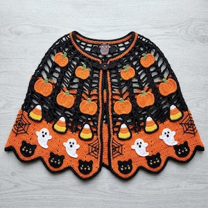 May include: A black and orange crocheted cape featuring a web-like pattern adorned with appliqués of pumpkins, candy corn, white ghosts, and black cats. The garment has a scalloped hem and a small owl-shaped button closure at the neckline.