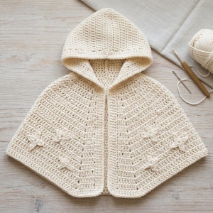 May include: A cream-colored hooded capelet crafted with a crochet stitch pattern. The garment features a pointed hood and a series of small, raised star-shaped embellishments integrated into the fabric across the front panels of the piece.