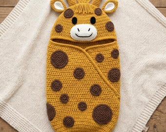 May include: A mustard yellow crochet baby swaddle blanket shaped like a giraffe. The hood features two small ears, two horns, and a tuft of yarn hair. The body is decorated with dark brown circular patches, and the face has a white muzzle.