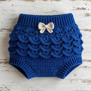 May include: A royal blue crochet diaper cover featuring a ribbed waistband and leg openings. The front is decorated with three horizontal rows of scalloped crochet shells and a small cream-colored bow with a central white pearl-like bead accent.