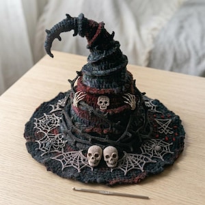 Op de afbeelding: Een donkere, gehaakte heksenhoed met een puntige, gebogen bovenkant, versierd met drie kleine schedels, skeletachtige handaccenten en ingewikkelde spinnenwebpatronen op de brede rand. Een zilverkleurige metalen haaknaald ligt voor de hoed.