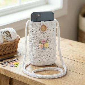 May include: A cream-colored crochet crossbody phone pouch featuring a leaf-like pattern, a wooden button closure, and a small cluster of pastel-colored fabric flowers. The pouch is accented with small white beads along the sides and has a long strap.