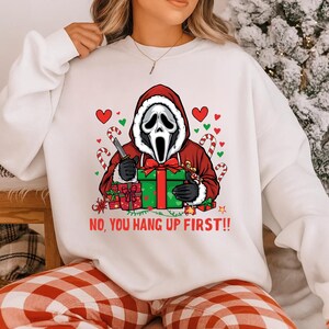 Könnte beinhalten: Weißes Sweatshirt mit einem Grafikdesign eines Scream-Maskenmannes im Weihnachtsmann-Kostüm, der ein Messer und ein Geschenk hält. Der Text "NO, YOU HANG UP FIRST!!" steht darunter. Zuckerstangen und Herzen umgeben die Figur.