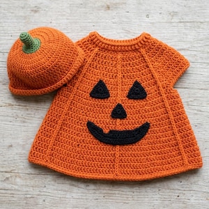 Puede incluir: Un vestido de ganchillo naranja brillante con un gorro a juego. El vestido presenta una cara de calabaza de Halloween tejida en negro con dos ojos triangulares, una nariz triangular y una sonrisa ancha y dentada. El gorro tiene un tallo verde.