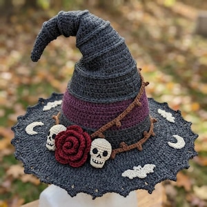 May include: A crocheted witch hat featuring a dark gray pointed crown and a wide brim. The hat is decorated with a deep red crocheted rose, two small cream-colored skulls, a brown vine-like trim, and white crocheted moon and bat motifs.
