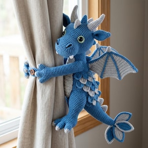 Op de afbeelding: Een gehaakte draak gordijnophouder in blauw en lichtgrijs met een textuur van schubben, twee vleugels, een lange staart en een bruine knoopsluiting op de handen om de stof vast te zetten. De draak heeft witte hoorns en kleine witte tanden.
