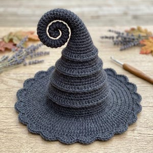 May include: A dark gray crocheted hat with a tall, spiraled, pointed crown and a wide, scalloped brim. The hat is constructed from yarn with a visible stitch pattern, featuring a distinct curled tip at the top of the cone-shaped structure.