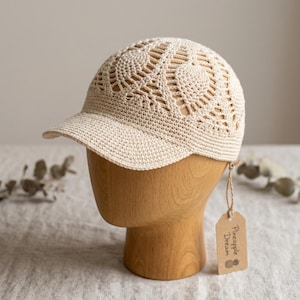 May include: A beige crochet baseball cap featuring an intricate openwork pattern across the crown and a solid brim. A small, light brown rectangular paper tag hangs from the side, printed with the text Pineapple Dream and a pineapple icon.