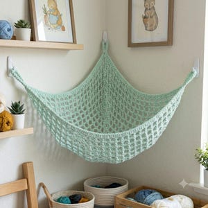 May include: A mint green crocheted corner hammock made of thick yarn with a diamond-shaped mesh pattern. The hammock is suspended by three white adhesive hooks attached to the walls, creating a storage space for soft items in a corner.