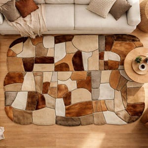 May include: Overhead view of a living room with a large, irregularly shaped rug in shades of brown, beige, and cream. The rug is the focal point, surrounded by a white sofa, a wooden coffee table, and a brown armchair. Decorative pillows and plants add to the cozy atmosphere.