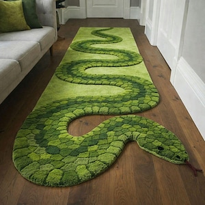 Puede incluir: Una alfombra verde con forma de serpiente, con un patrón texturizado en forma de escamas en diferentes tonos de verde. La alfombra está colocada sobre un suelo de madera, con la cabeza y la cola de la serpiente visibles. Un artículo de decoración único.