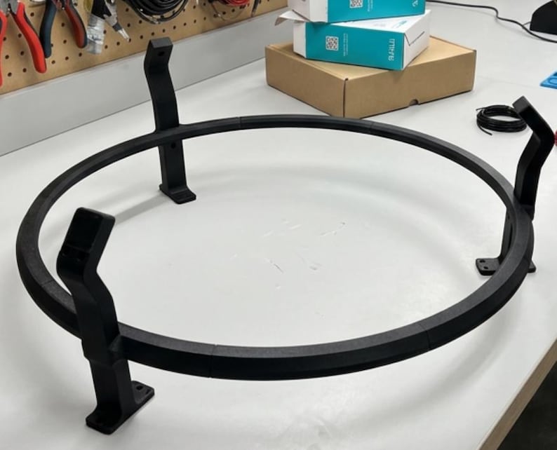 Puede incluir: Un soporte circular de metal negro con tres patas de soporte. El soporte tiene una parte superior redonda y est&aacute; dise&ntilde;ado para sostener un objeto. El soporte es de metal y tiene un acabado mate.