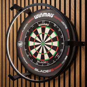 Puede incluir: Un tablero de dardos Winmau Blade 6 negro y rojo con un dise&ntilde;o de diana verde y blanco. El tablero de dardos est&aacute; montado en una pared de madera con un marco de metal negro y una luz circular.