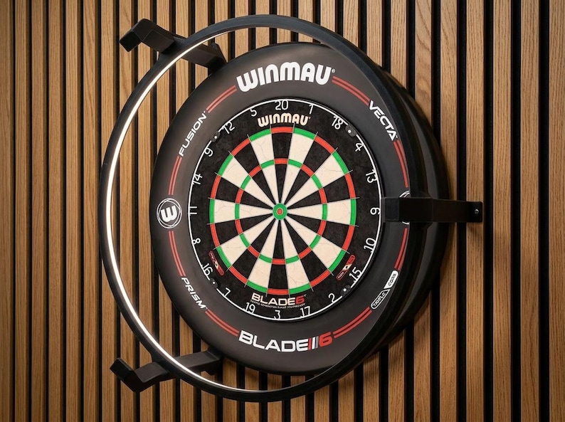 Puede incluir: Un tablero de dardos Winmau Blade 6 negro y rojo con un dise&ntilde;o de diana verde y blanco. El tablero de dardos est&aacute; montado en una pared de madera con un marco de metal negro y una luz circular.
