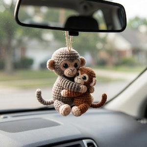 May include: A crocheted hanging ornament featuring two monkeys, one larger in light gray and one smaller in brown, embracing each other. The monkeys have dark plastic eyes, stitched facial features, and long tails, suspended by a thin twine string.