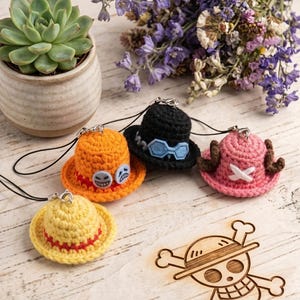 May include: Four miniature crocheted hats with black attachment cords. One yellow hat with a red band, one orange hat with two circular face icons, one black hat with blue goggles, and one pink hat with brown antlers and a white cross emblem.