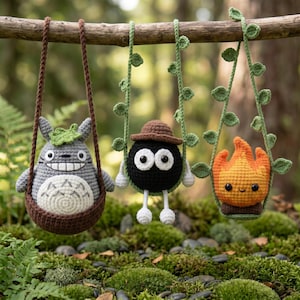 May include: Three crocheted plush figures hanging from a branch. A gray creature with a white belly and leaf on its head, a black round soot sprite wearing a brown hat, and an orange flame character, each suspended by crocheted vine straps.