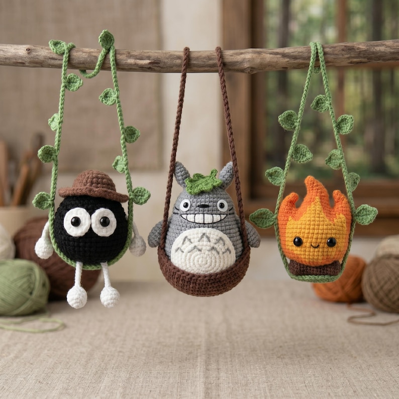 May include: Three crocheted hanging ornaments suspended from a wooden branch. The left is a black sphere with a brown hat, the center is a gray creature with a white belly and green leaf, and the right is an orange flame shape with a smiling face.