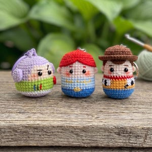 May include: Three crochet keychains featuring stylized characters. The left is purple, green, and white. The center has red hair, a blue and white shirt, and a yellow collar. The right wears a brown hat, a yellow and red shirt, and blue pants.
