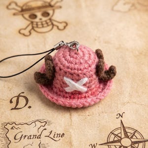 May include: A small, pink, crocheted hat charm featuring two brown antlers on the sides and a white stitched X symbol on the front. The item includes a black cord and a silver metal lobster clasp for attachment, designed as a miniature accessory.