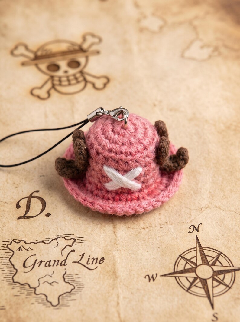 May include: A small, pink, crocheted hat charm featuring two brown antlers on the sides and a white stitched X symbol on the front. The item includes a black cord and a silver metal lobster clasp for attachment, designed as a miniature accessory.