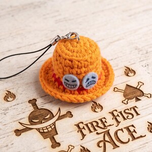 May include: A miniature orange crochet hat charm featuring two small circular felt patches with embroidered faces. The hat has a red band and is attached to a black cord with a silver metal lobster clasp. The design is inspired by the character Fire Fist Ace.