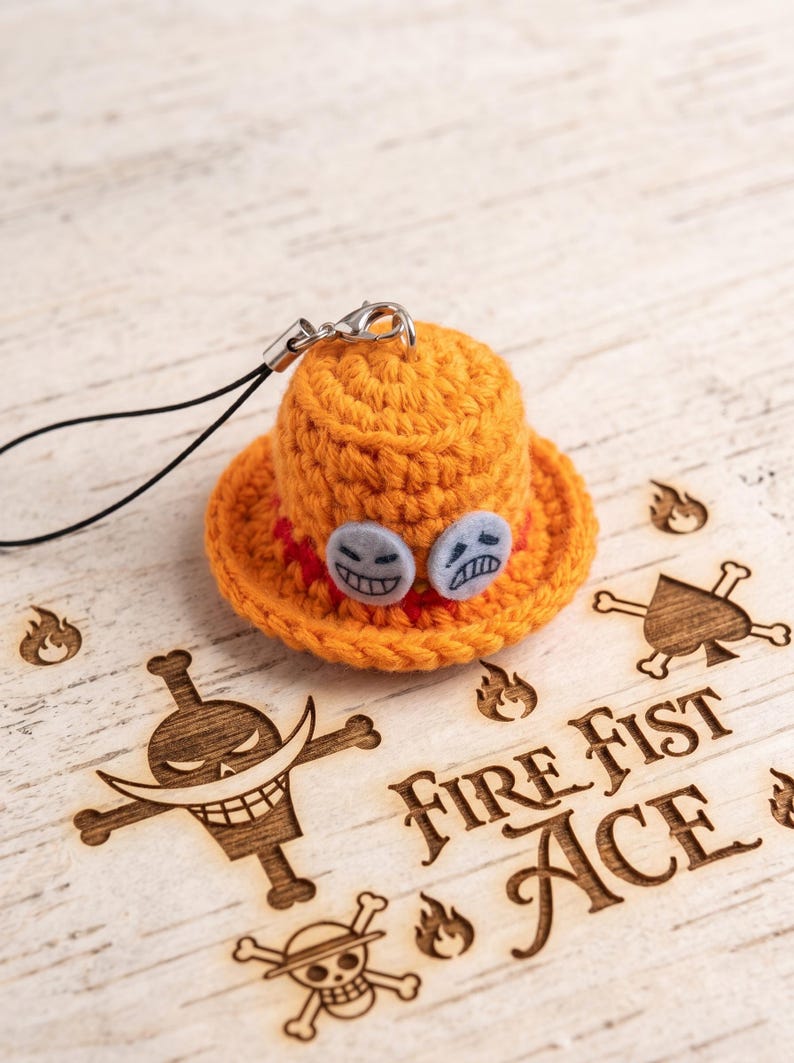 May include: A miniature orange crochet hat charm featuring two small circular felt patches with embroidered faces. The hat has a red band and is attached to a black cord with a silver metal lobster clasp. The design is inspired by the character Fire Fist Ace.