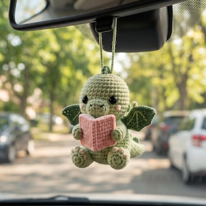 May include: A small, light green crocheted dragon hanging ornament featuring dark eyes, pink cheeks, and small wings. The dragon is holding an open, light pink crocheted book in its hands, suspended by a matching green yarn loop for display.