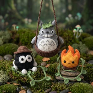 May include: Three crocheted figures: a gray creature with a white belly and leaf on its head hanging in a brown basket, a black round creature wearing a brown hat, and an orange flame-shaped creature with a smiling face and green vines.