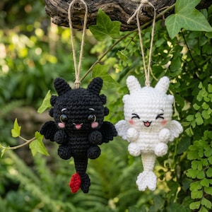 May include: Two crocheted dragon ornaments hanging from twine. One is black with large dark eyes, pink cheeks, and a red tail tip. The other is white with large dark eyes, pink cheeks, and a white tail. Both have small wings and ears.