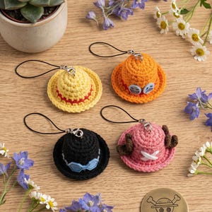 May include: Four miniature crocheted hats with black phone straps. One yellow hat with a red band, one orange hat with blue button accents, one black hat with blue goggles, and one pink hat with brown antlers and a white cross emblem.