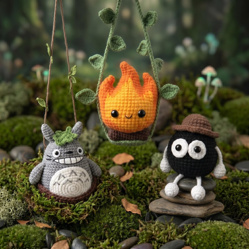 May include: Three crocheted figures: a gray Totoro with a white belly, an orange and yellow flame character suspended in a green vine swing, and a black soot sprite wearing a brown hat, all crafted with detailed yarn textures and stitching.