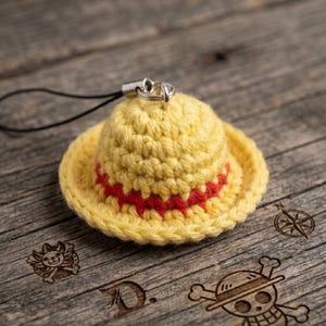 May include: A miniature yellow crocheted straw hat charm featuring a red band around the crown. The hat is attached to a black cord loop with a silver-toned metal jump ring. The design mimics a classic sun hat style with a rounded top and brim.