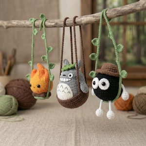 May include: Three crocheted figures hanging from a wooden branch: an orange flame character on a green vine swing, a gray Totoro-like creature in a brown basket, and a black soot sprite with a brown hat and white limbs on a green vine swing.