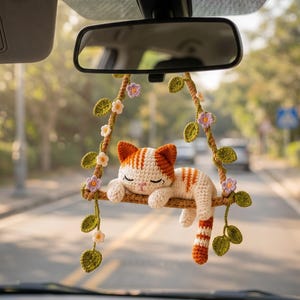 May include: A crocheted car accessory featuring a sleeping orange and white tabby cat resting on a brown swing. The swing is decorated with green crocheted leaves and small purple and white flowers, suspended by brown yarn ropes from a rearview mirror.