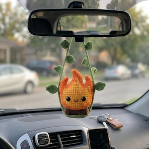 May include: A crocheted hanging ornament featuring a small, smiling flame character with orange and yellow yarn, sitting atop a brown base. The flame is framed by two green crocheted vine strands with small leaves, designed to hang from a rearview mirror.