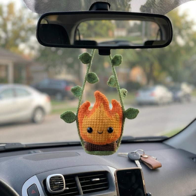 May include: A crocheted hanging ornament featuring a small, smiling flame character with orange and yellow yarn, sitting atop a brown base. The flame is framed by two green crocheted vine strands with small leaves, designed to hang from a rearview mirror.