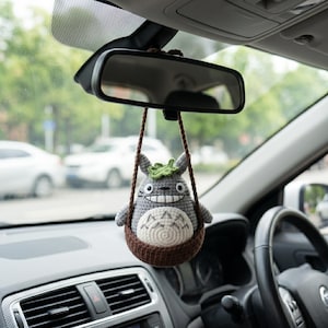 May include: A gray and white crocheted figure with pointed ears and a green leaf on its head, sitting inside a brown crocheted basket. The figure has a wide, toothy grin and dark eyes, and it hangs from a brown cord attached to a car rearview mirror.