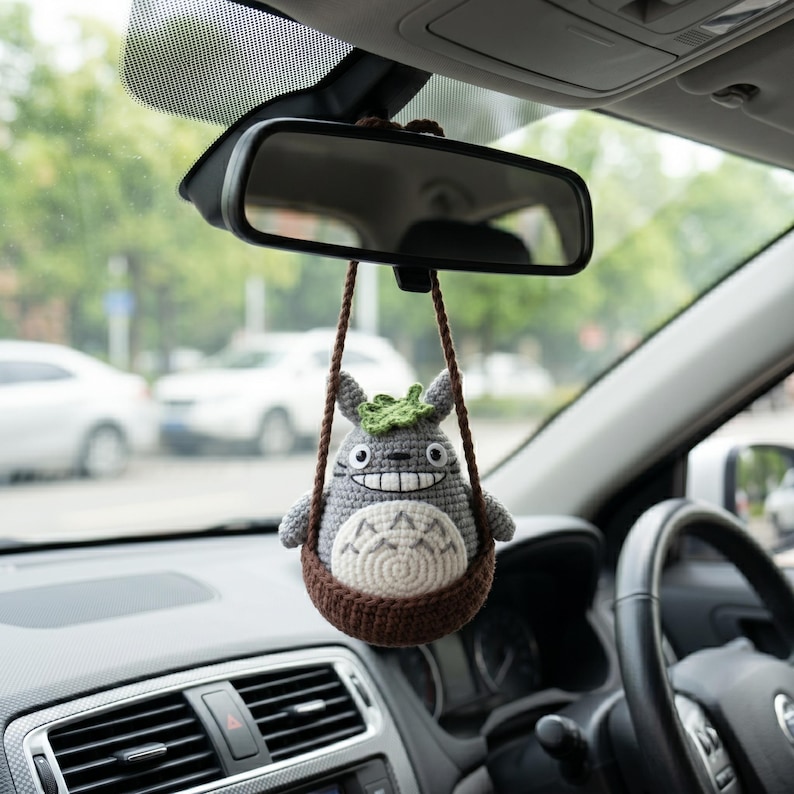 May include: A gray and white crocheted figure with pointed ears and a green leaf on its head, sitting inside a brown crocheted basket. The figure has a wide, toothy grin and dark eyes, and it hangs from a brown cord attached to a car rearview mirror.