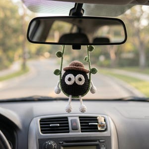 May include: A crocheted black soot sprite ornament with large white eyes, wearing a brown hat, hanging from a green vine-like cord. The round figure has small white hands and feet, suspended from a car rearview mirror in a vehicle interior.