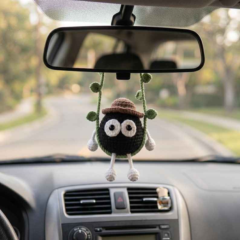 May include: A crocheted black soot sprite ornament with large white eyes, wearing a brown hat, hanging from a green vine-like cord. The round figure has small white hands and feet, suspended from a car rearview mirror in a vehicle interior.
