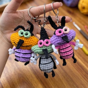 May include: Three handmade crochet keychains in the shape of cartoon insects. The keychains feature various colors, including orange, green, and purple, with black and white accents. Each keychain has a silver keyring attached.