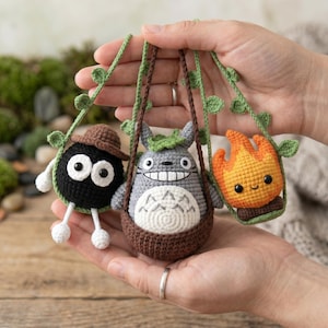 May include: Three small crocheted figures hanging from a green vine-like cord. One is a black round creature with a brown hat, one is a gray creature with a white belly and leaf on its head, and one is an orange flame-shaped figure with a smiling face.