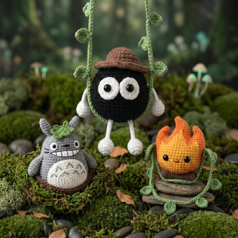 May include: Three crocheted figures: a round black soot sprite with a brown hat hanging from green vines, a grey Totoro-like creature with a white belly and leaf on its head, and an orange flame character sitting on a stack of grey stones.